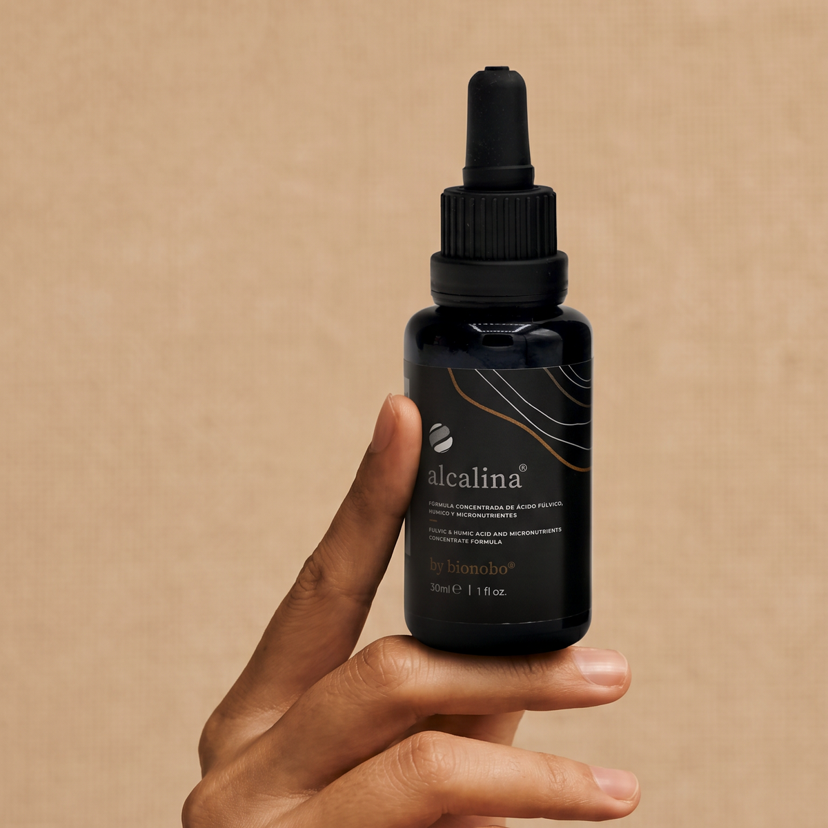 A raised hand holding the alcalina dropper bottle against a warm pink-taupe wall, the label centred and legible.