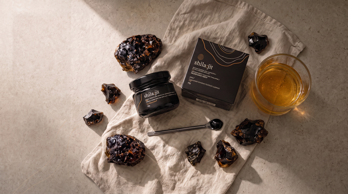 Open jar of Bionobo golden-grade Shilajit resin with its carton, mineral rocks, measuring spoon and a glass of water.